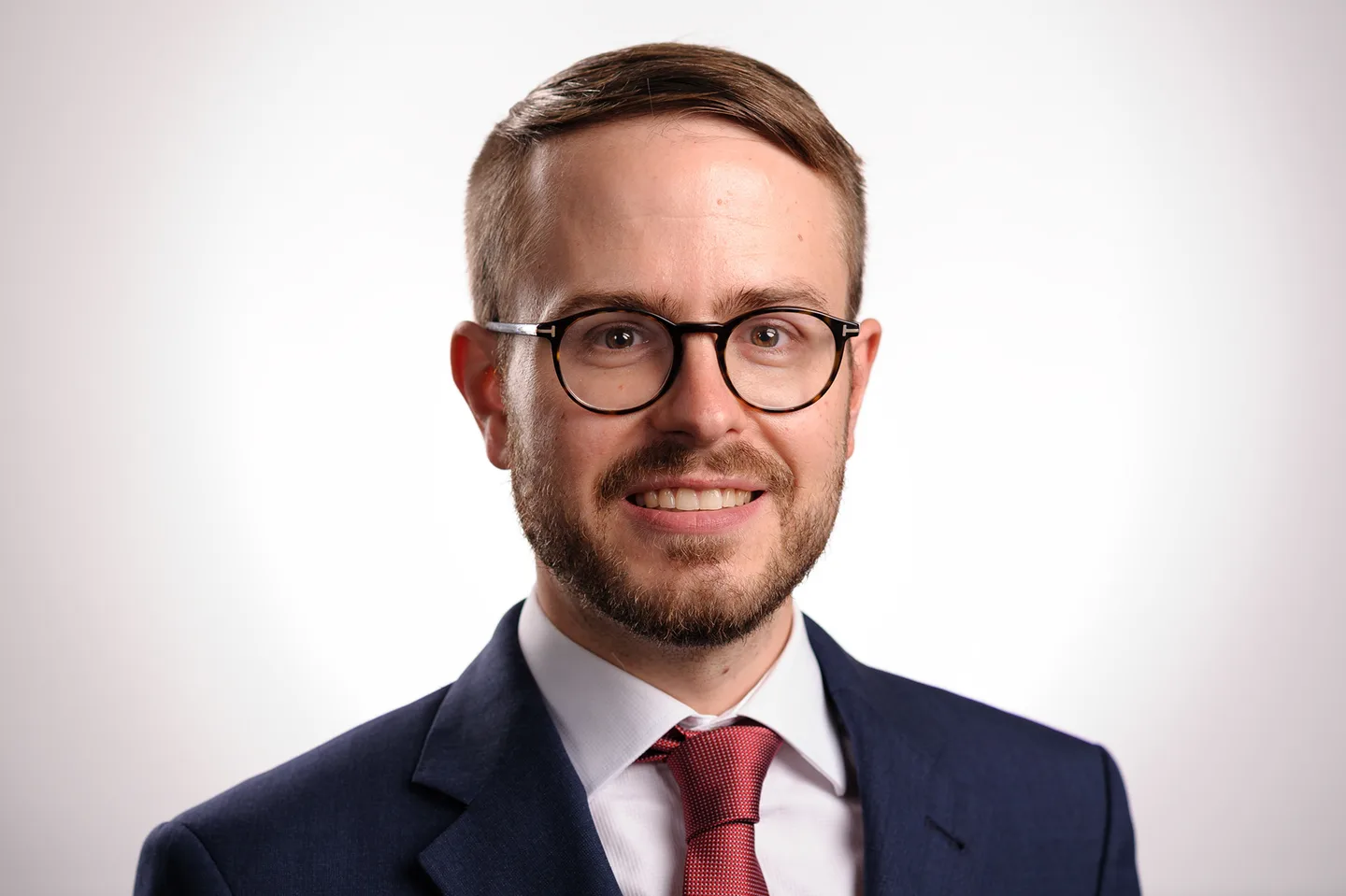 Studio headshot of a smiling man with round glasses and a short beard in a navy suit and red tie