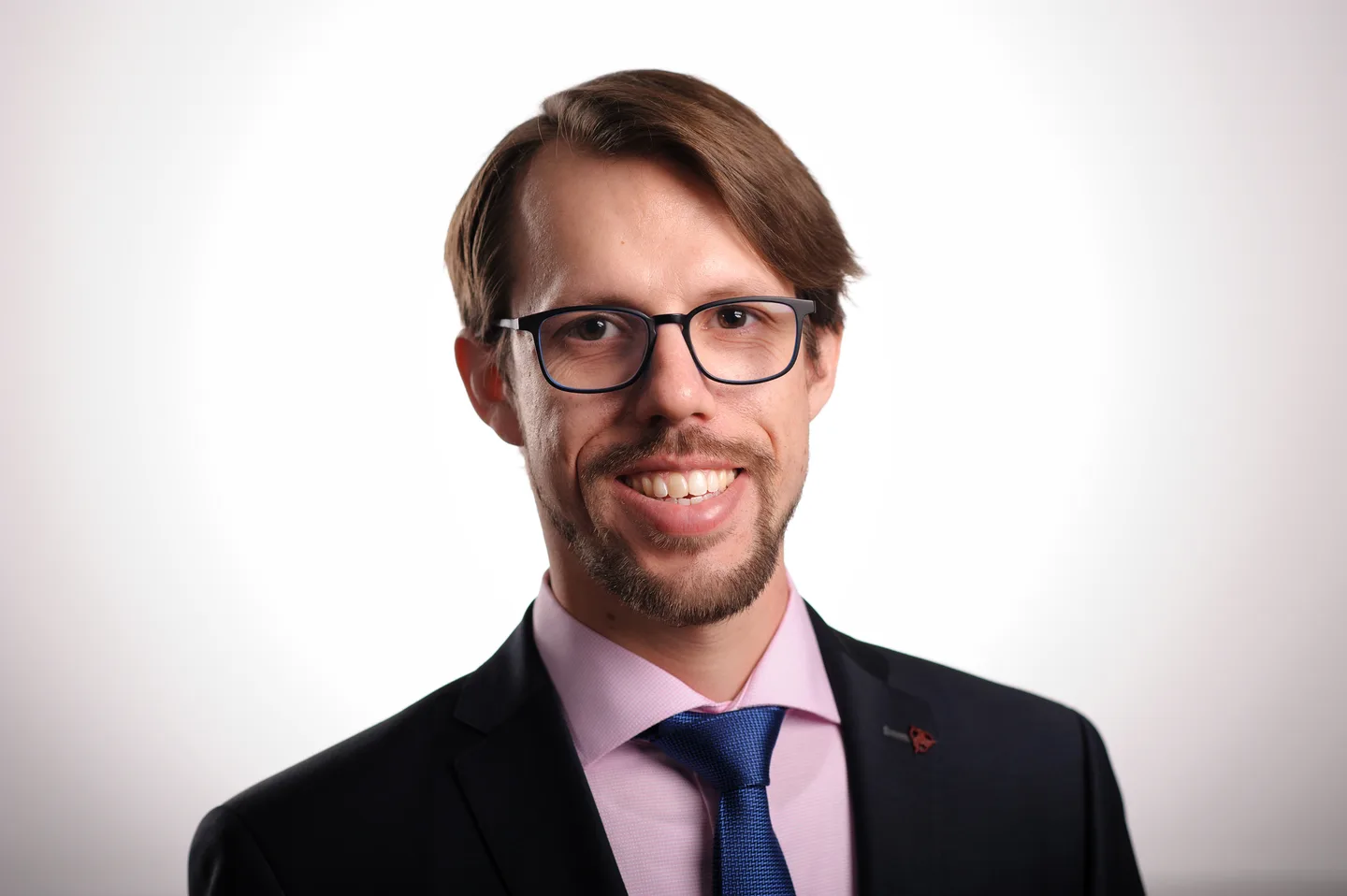 Studio headshot of a laughing man with glasses and a goatee in a dark suit, pink shirt and blue tie